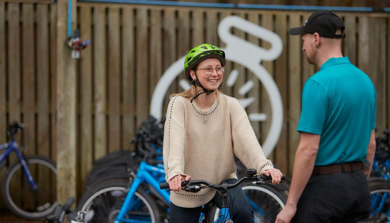 Cyclist wearing a green helmet smiles while holding the handlebars, conversing with a staff member. Surroundings include blue bicycles, stacked tires, and a wooden fence with a large white logo.