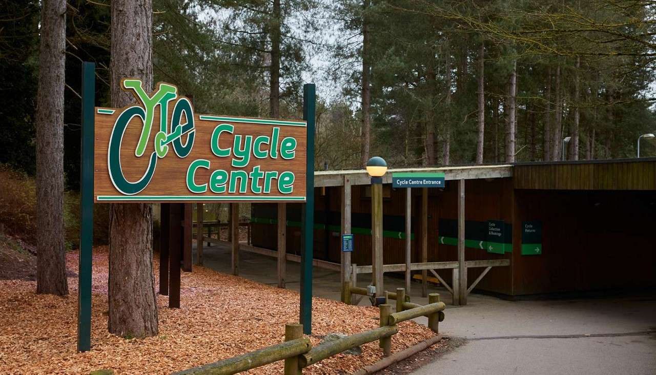 Cycle Centre sign stands, directing visitors; a covered walkway leads through a wooded area. Visible text: “Cycle Centre,” “Cycle Centre Entrance,” “Cycle Collection & Bookings,” “Cycle Returns.”