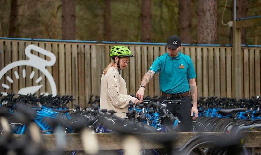 Staff member points at a bicycle while a helmeted customer watches; rows of bikes surround them in a fenced outdoor rental area amid tall forest trees.