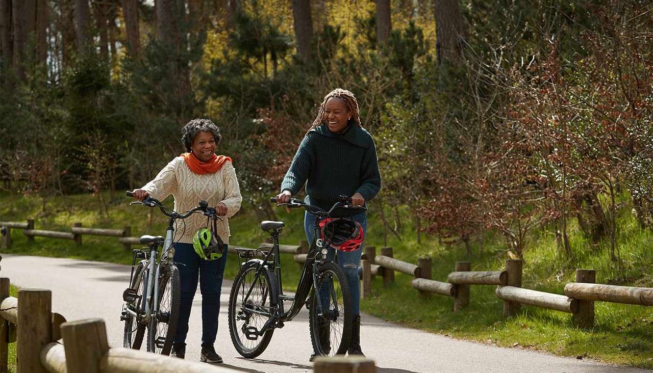 Two people push bicycles while chatting, helmets hanging from handlebars, on a paved path bordered by wooden rails amid green grass, shrubs, and tall trees in soft sunlight.