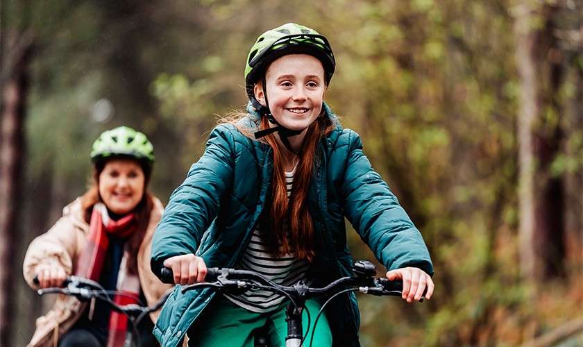 A helmeted person rides a bicycle, smiling, hands on handlebars; another cyclist follows behind; context: wooded trail with soft light and blurred trees.
