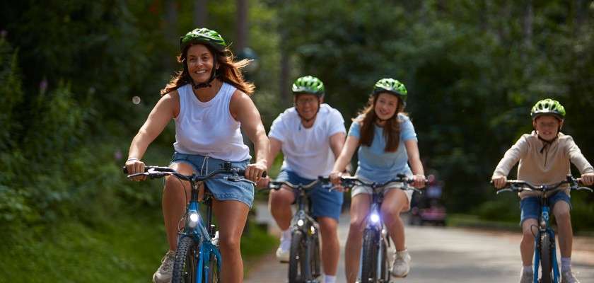 Four bicyclists ride forward, smiling and wearing helmets, on a paved path; surrounding trees and greenery indicate a wooded park setting on a sunny day.