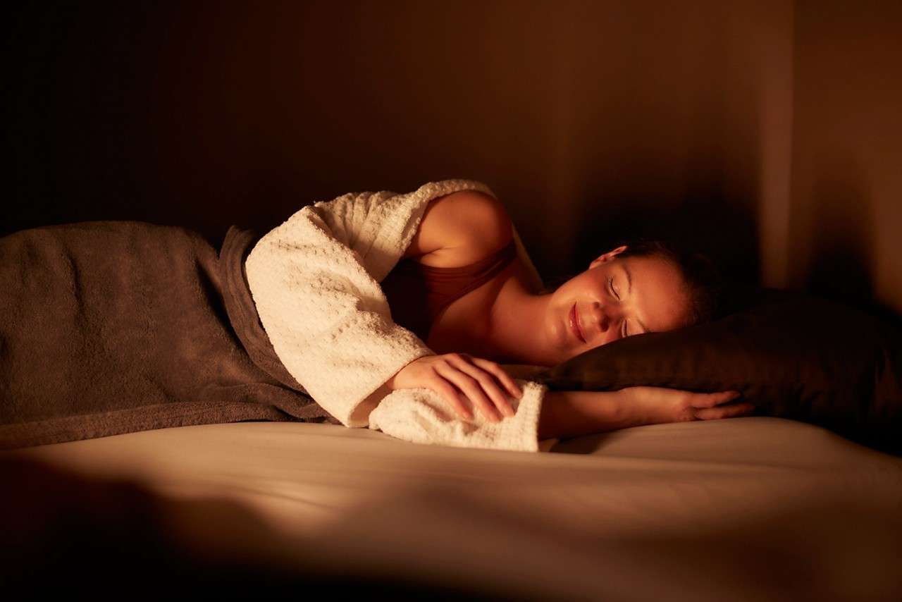 Person sleeps on their side, smiling slightly, head on dark pillow. Warm lamplight fills a dim bedroom. They wear a robe and blanket draped over them on a bed.