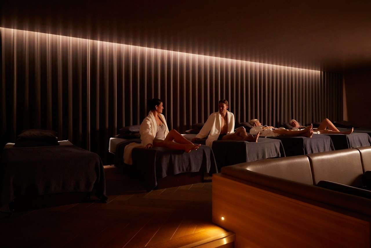 Spa guests lounge and chat on padded daybeds while others recline resting. Warm, low lighting and ribbed wall panels create a calm, dim relaxation-room ambiance.