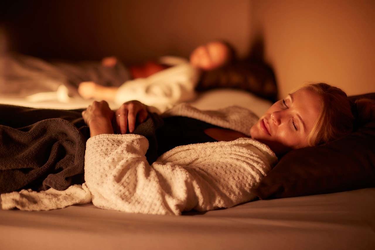 Woman sleeps on a cushioned bed, hands clasping a dark blanket, wearing a white robe; another person rests blurred in background within a warm, dimly lit spa-like relaxation room.