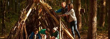 Stick shelter; adults position branches while children crouch inside; in a sunlit forest among tall trees and leaf litter, suggesting outdoor play or survival activity.