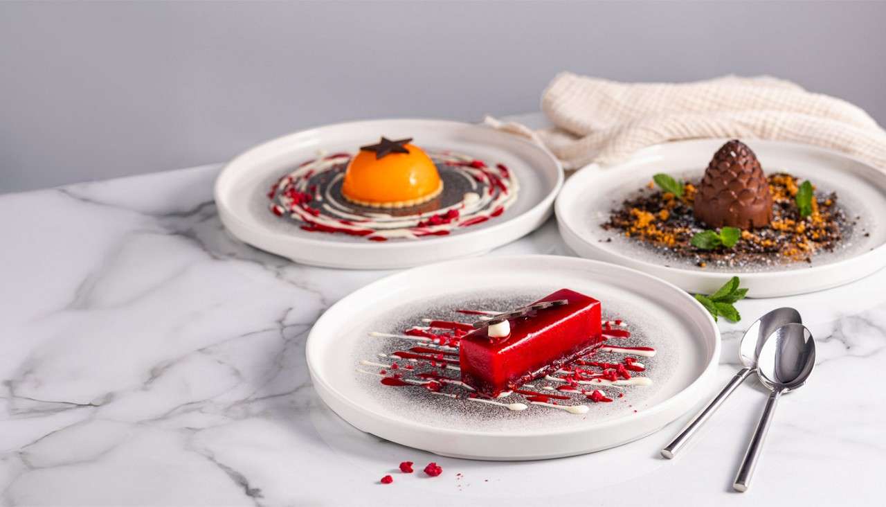 Three plated desserts sit artfully arranged; a glossy red rectangular mousse drizzled with sauces, an orange dome tart, and a chocolate pinecone, on white plates atop a marble countertop with spoons.