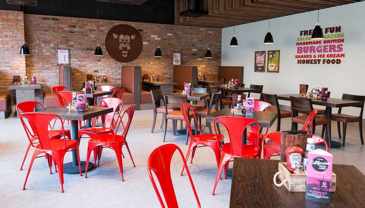 Restaurant seating—red metal chairs surround wooden tables; brown booths line brick and pale walls under black pendant lights in a casual diner.

Text:
- FRESH FUN
- SALAD [unreadable]
- HANDMADE BRITISH
- BURGERS
- SHAKES & ICE CREAM
- HONEST FOOD
- BB (logo)
- Heinz Tomato Ketchup
- HP Sauce
- California Fresh
- Additional small poster and menu text unreadable