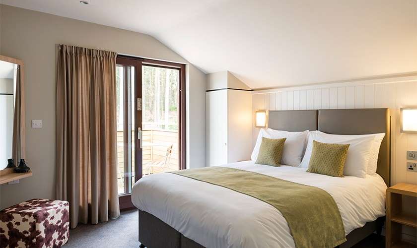 Bed sits neatly made with white linens and green accents, in a cozy modern bedroom; bedside lamps glow, curtains frame glass door opening to a wooded-view balcony with chairs.