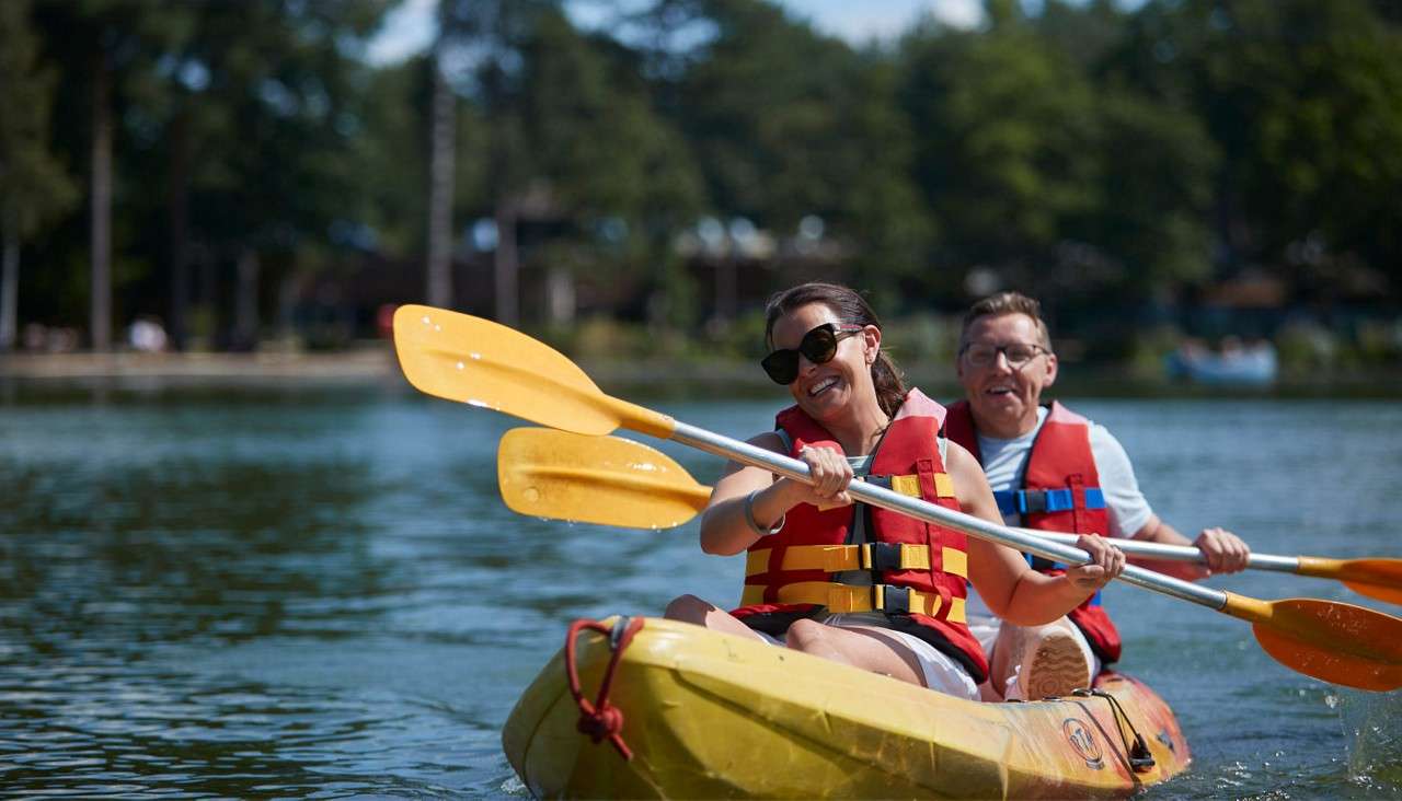 Two adults in a yellow tandem kayak paddle, smiling, wearing red life vests; water splashes as they cross a calm lake, with blurred trees and shoreline in the background.