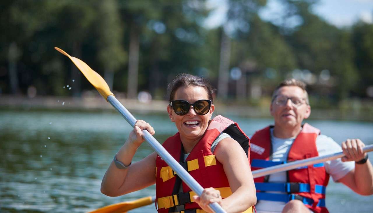 Two adults in red life vests paddle a kayak, front person smiling and splashing water, while the rear steers; on a calm lake surrounded by trees on a sunny day.
