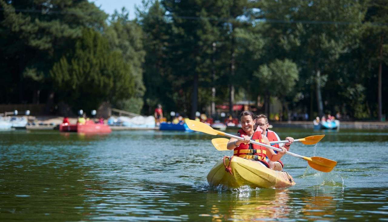 Two people in a yellow kayak paddle forward, splashing water. They wear life vests. In the background, other boats and a dock sit on a tree-lined lake under clear daylight.
