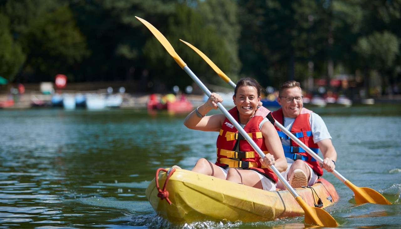 Two kayakers paddle a yellow tandem kayak, smiling and splashing, wearing red life jackets on a calm lake; trees and moored boats in the background. Visible text: “JOBE,” “S”.