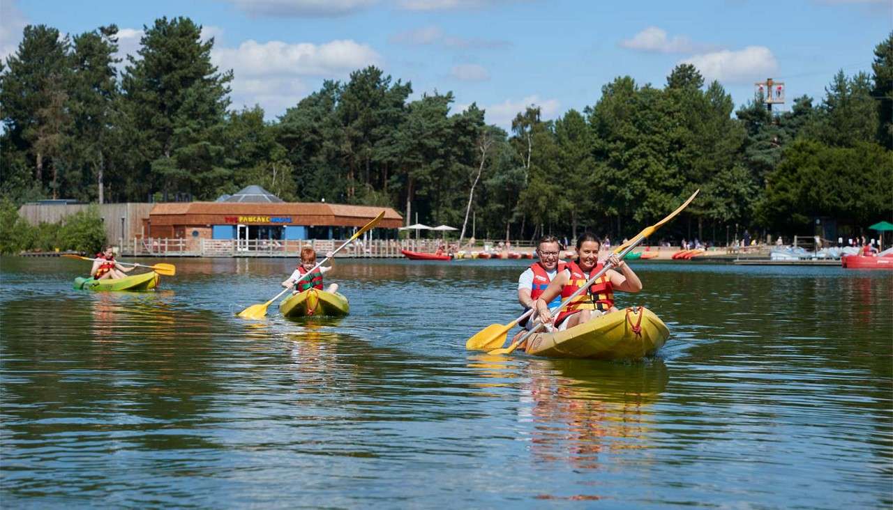 Kayakers paddle yellow boats forward, splashing water. In the background, a lakeside building and dock sit among tall trees under blue sky; its sign reads: THE PANCAKE HOUSE.