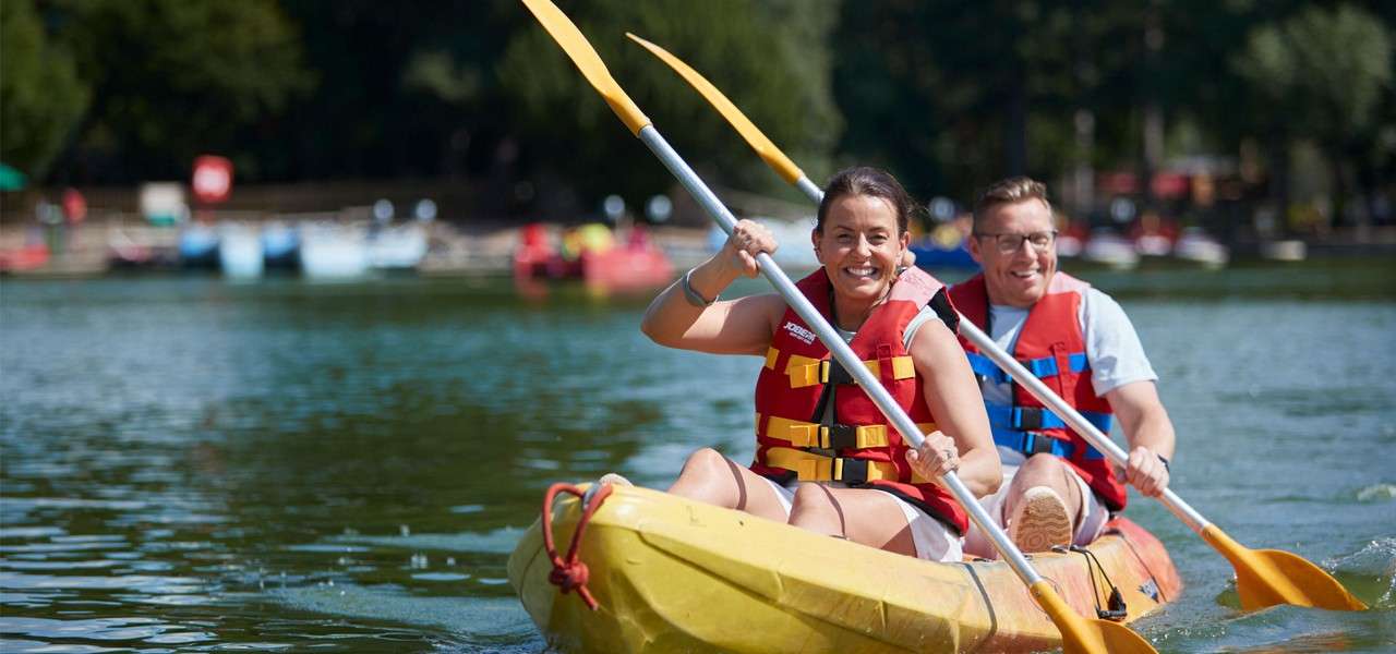 Two adults in a yellow tandem kayak paddle vigorously, smiling, wearing red life jackets, on a calm lake; trees and docked boats blur in background. Visible text: JOBE.