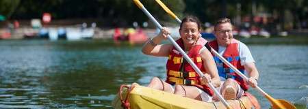 Two people in a yellow tandem kayak paddle forward, smiling, wearing red life vests; calm lake water splashes around them, with distant boats and trees in a sunlit park.