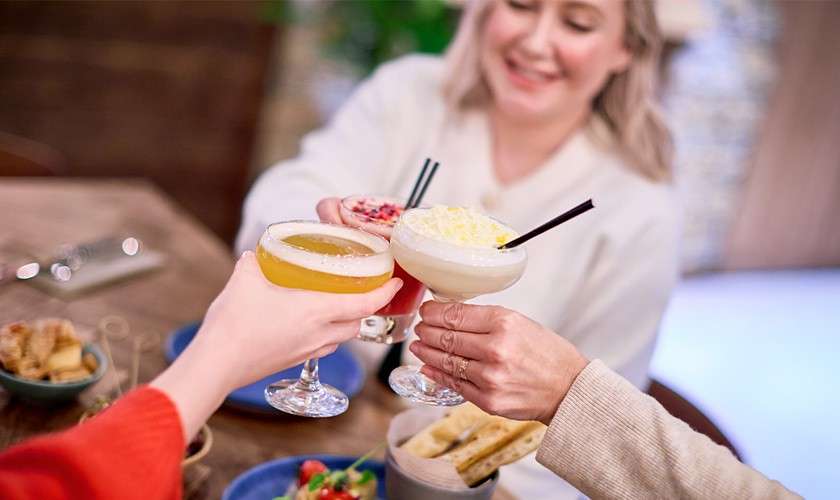 Three cocktail glasses toast, held by hands; drinks include a yellow sour and a white frozen. A diner sits at a table with small plates, breadsticks, and blurred restaurant background.