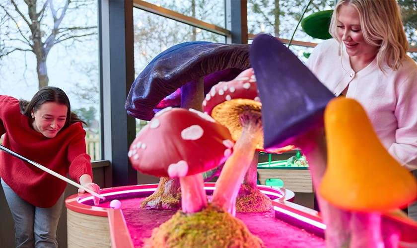 Mini-golf ball rolls as two people aim with a putter. Oversized, colorful mushroom obstacles glow with pink lighting. Indoor course setting with large windows revealing trees outside.