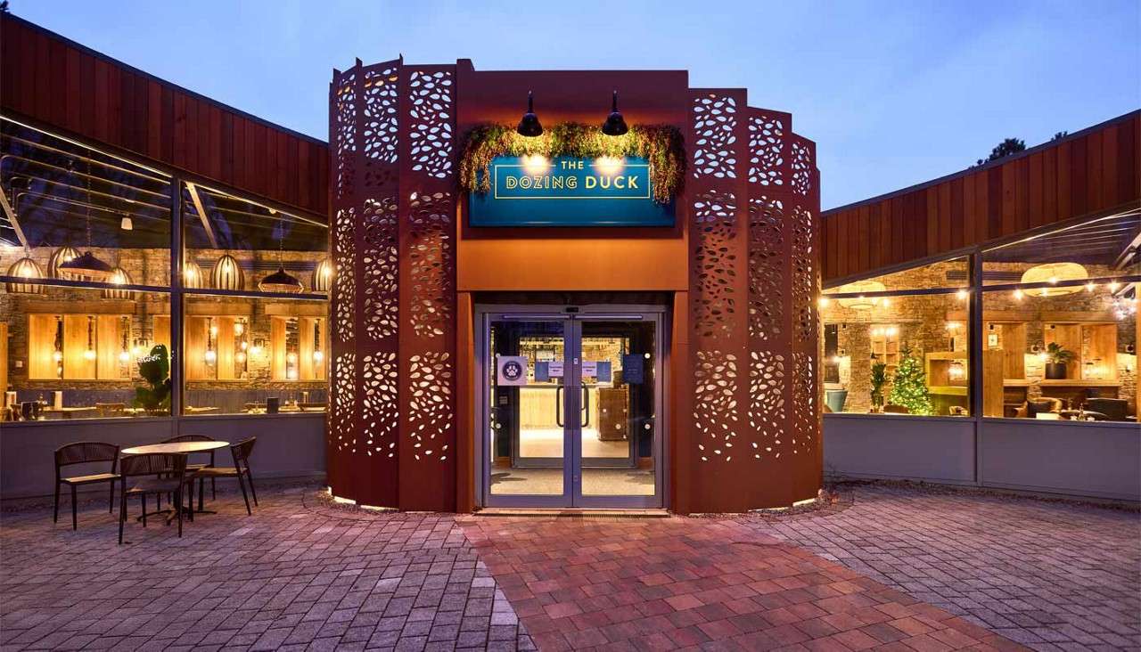 Restaurant entrance glows under lamps, displaying sign: “THE DOZING DUCK”; glass doors centered, perforated metal panels flank; context: evening patio with empty tables and warm, lit interior visible.
