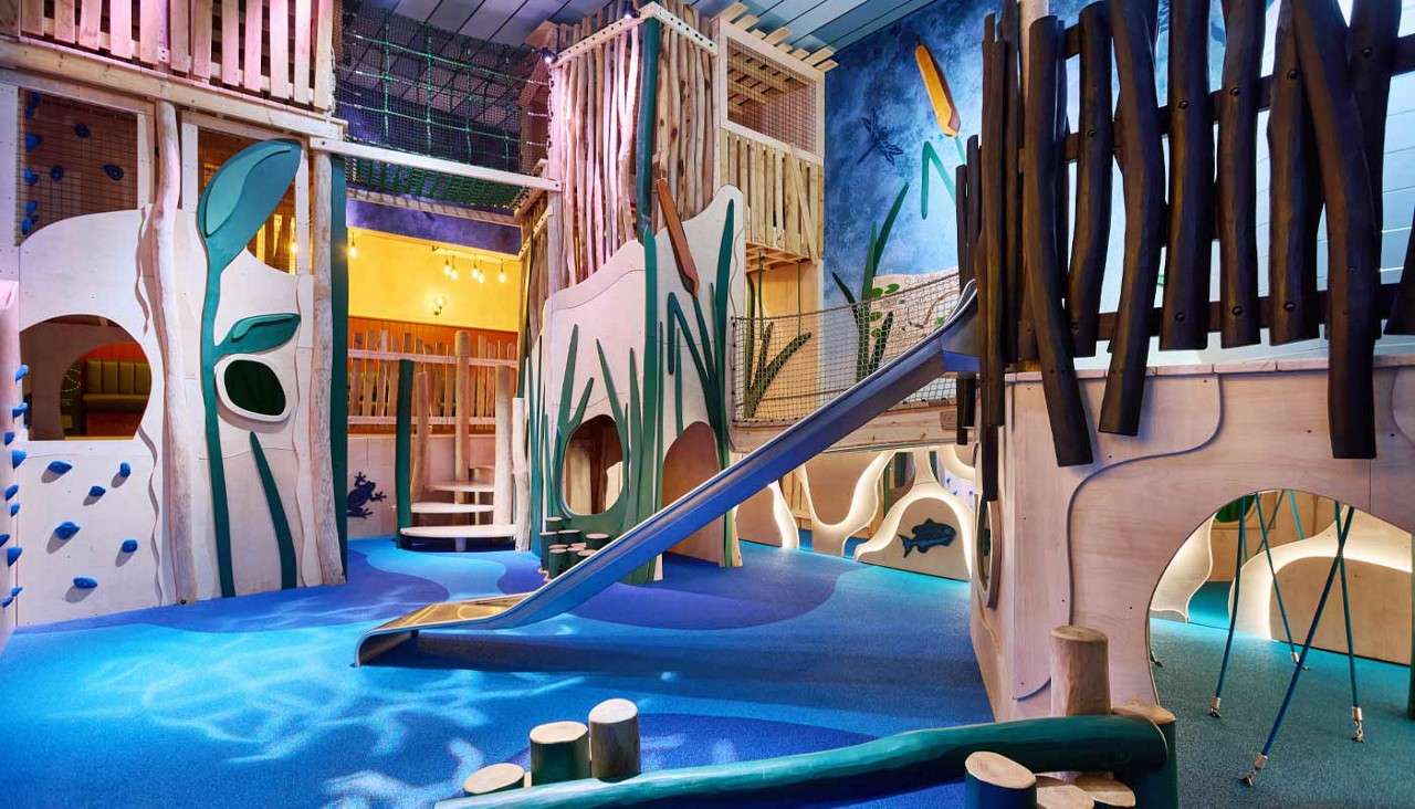Indoor wooden playground features a long metal slide descending from a bridge, with climbing walls, rope nets, tunnels, and lily-pad decor, set over blue cushioned flooring in a nature-themed room.