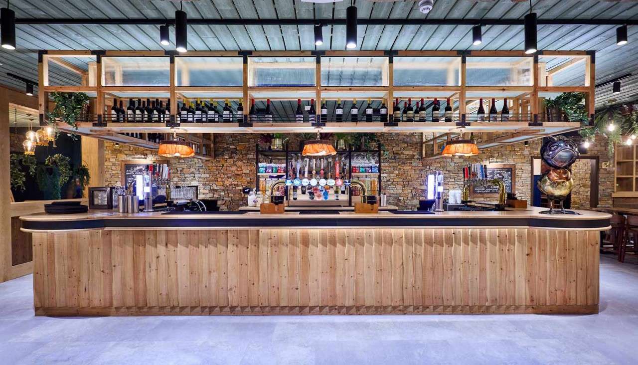 Wooden bar counter anchors the scene, displaying beer taps and bar tools, while overhead wine bottles and pendant lamps hang; set within a rustic pub with stone walls and greenery.