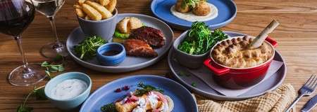 Assorted plated dishes rest on a wooden table: meat pie in red crock, fries, steak with sauce, greens, croquettes, seafood bites, dips, and two glasses of wine.