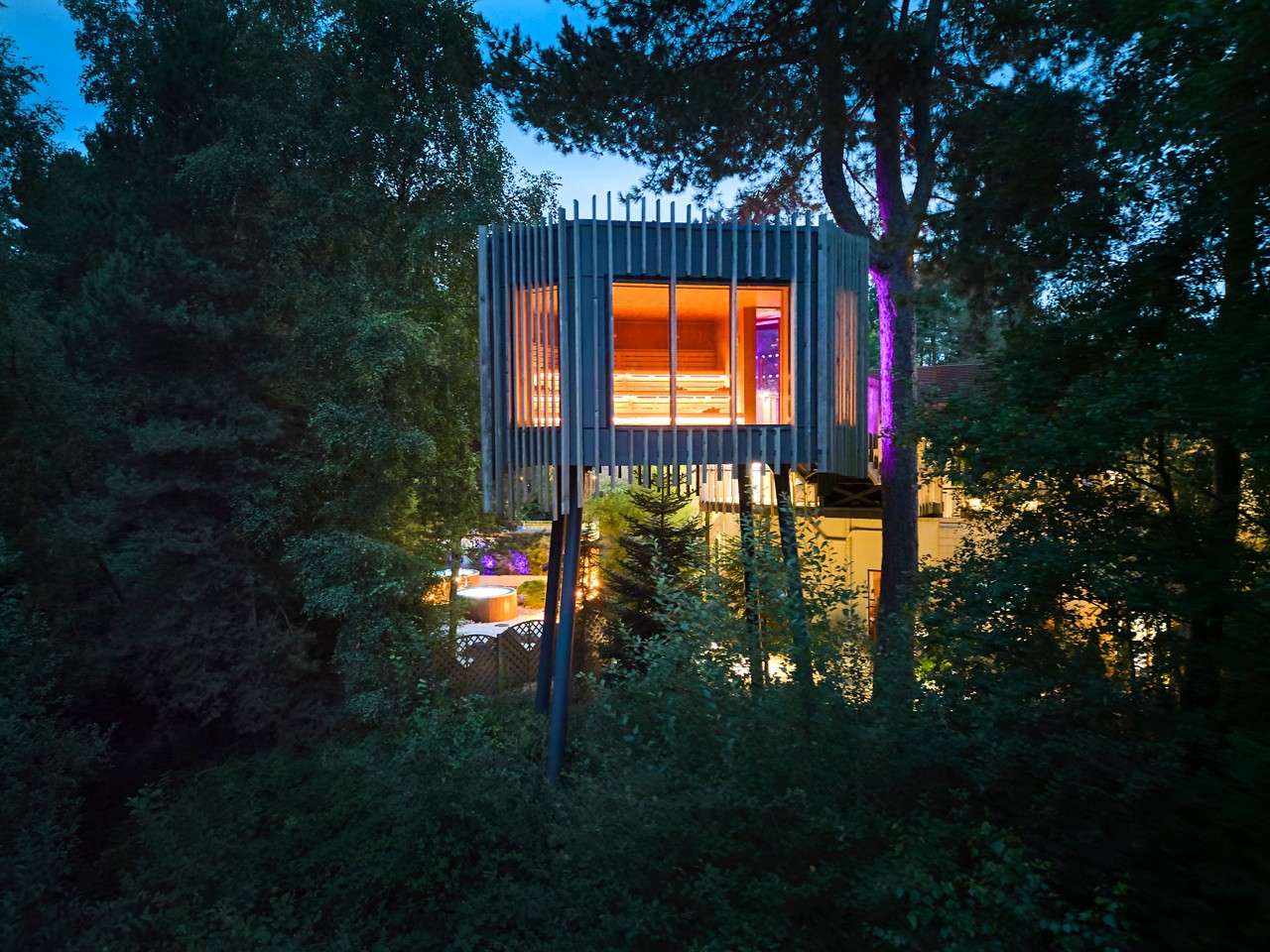 Modern treehouse glows with warm interior light, perched on tall stilts; large windows reveal wooden benches. Dense forest at dusk surrounds it, with nearby illuminated buildings and purple accent lighting.