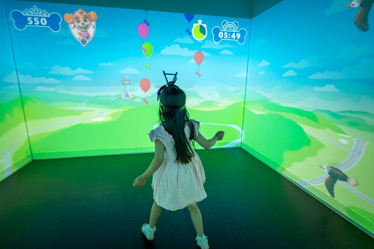 Child wearing a headband tracker moves toward floating balloons, playing an interactive projection game; animated hills surround; on-screen score and timer read "550" and "05:49".
