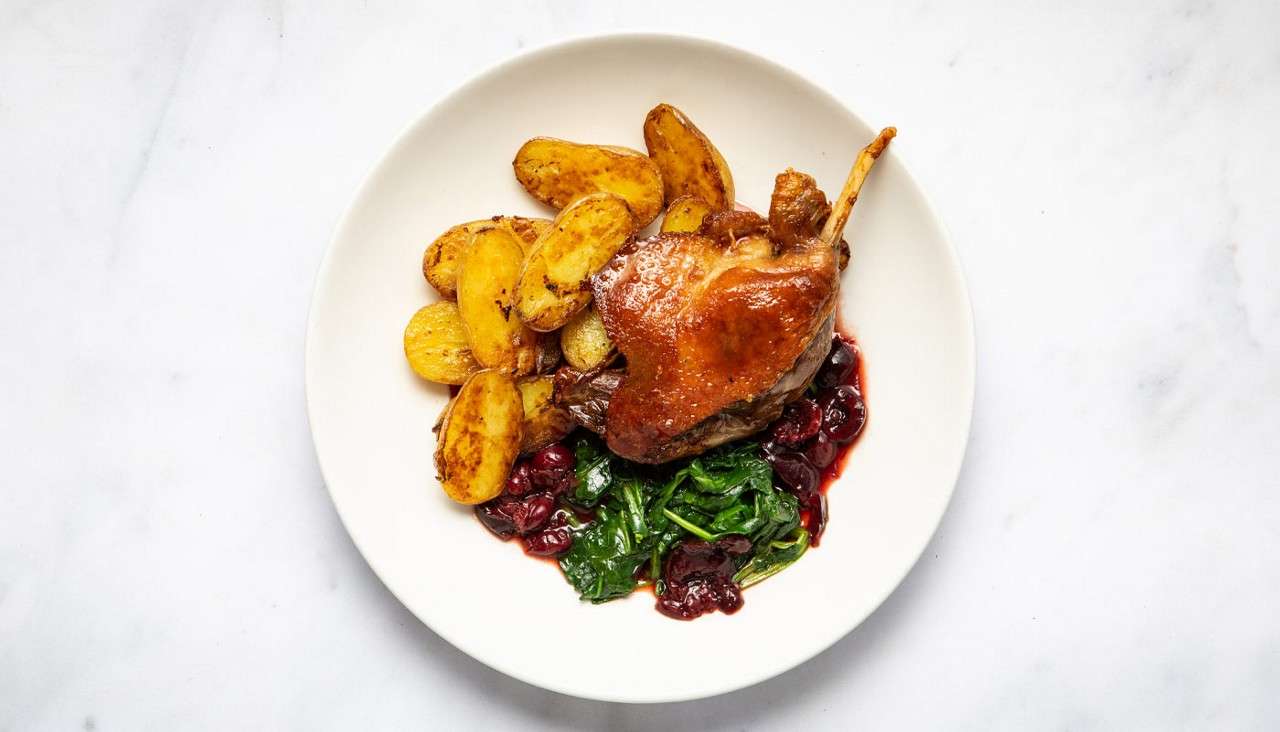 Roasted poultry leg rests beside sides; golden roasted potato halves and sautéed greens sit around, with a glossy dark red cherry-like sauce, all arranged on a white plate.