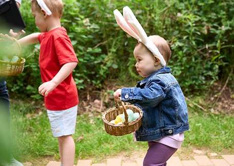 Animatronic reindeer heads sing with moving mouths, mounted in wooden stalls; a bundled-up man and two children watch from a railing inside a festive, light-trimmed shed with a woodchip floor.