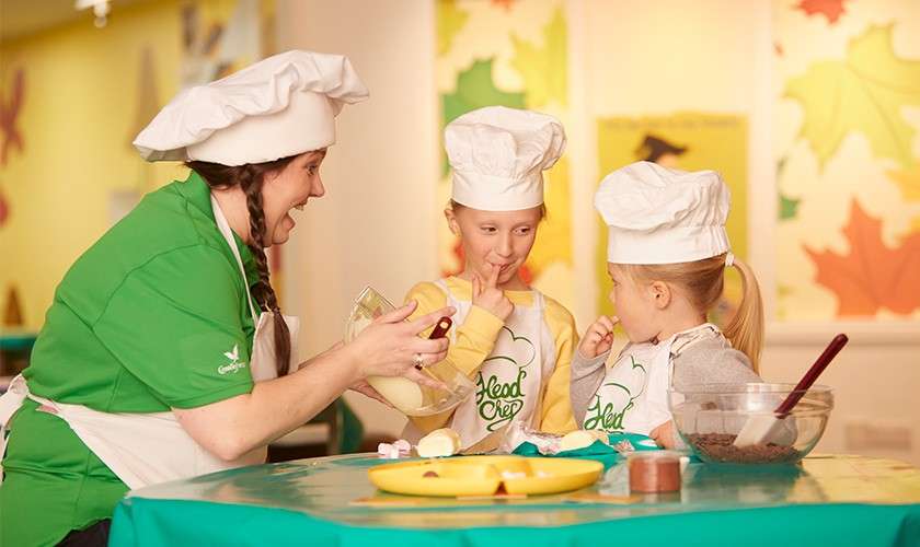 Two children in chef hats lick utensils as an adult pours batter from a bowl at a green-topped table with bowls and tools in a bright room. Text: “Head Chef.”