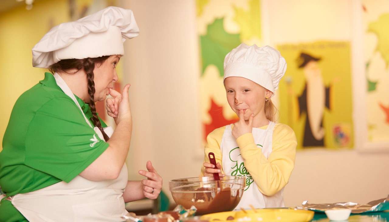 Two apron-wearing bakers lick chocolate from their fingers while mixing batter in a glass bowl with a spatula, in a brightly lit classroom kitchen decorated with colorful posters and scattered cooking supplies.