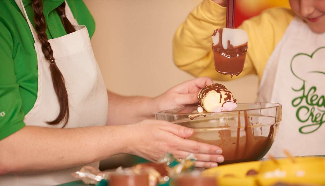 Dessert scoop with marshmallows is drizzled with melted chocolate, held above a glass bowl by two apron-wearing people in a kitchen. Visible text on a child's apron: Head Chef.
