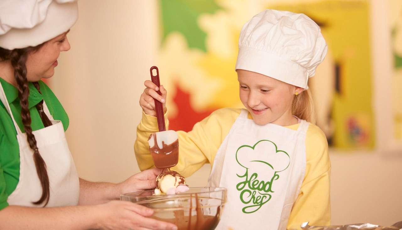 Child chef pours melted chocolate from a spatula onto an ice-cream scoop, assisted by an adult, over a mixing bowl; both wear white hats and aprons. Visible text: Head Chef.