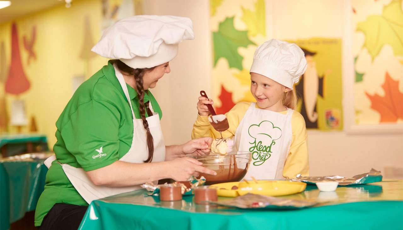 Child chef dips a marshmallow into melted chocolate while smiling at an instructor. Cooking-class setting with mixing bowl, utensils, and leaf-themed posters. Text: “Head Chef” (apron), “Center Parcs” (sleeve).