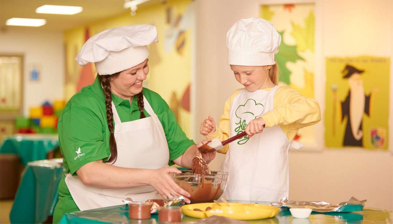 Two people wearing chef hats mix melted chocolate with a spatula in a glass bowl; the adult guides the child in a bright classroom with green tablecloths and colorful posters.
Text: “Center Parcs” (adult’s shirt).