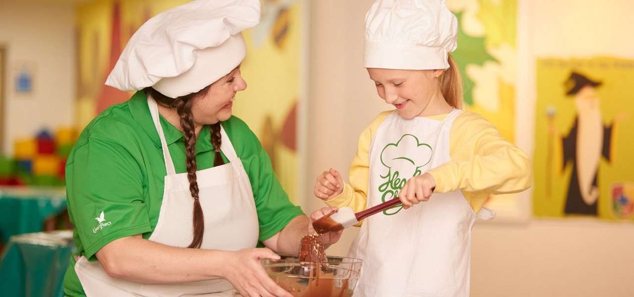 Child baker stirs chocolate batter with a spatula while an adult holds the bowl, both wearing chef hats and aprons in a bright classroom with colorful art. Text: Le Chef.