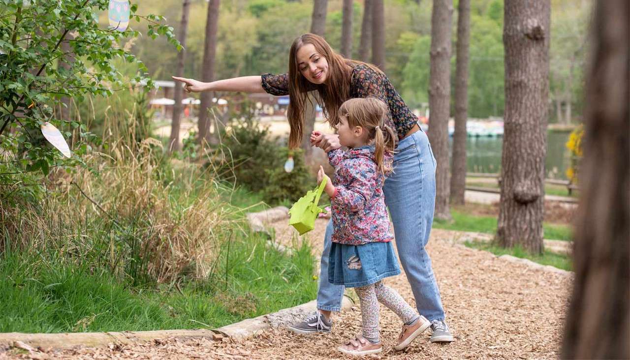 Woman points while a child holds a green basket, walking on a woodland path; paper egg decorations hang from a bush; pine trees and a lake appear in the background.