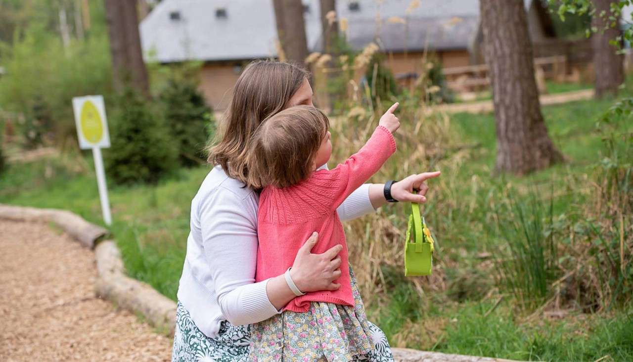Child in pink sweater points ahead while being held by an adult, both kneeling on a wooded path; adult points and holds a green bag near trees and cabins.