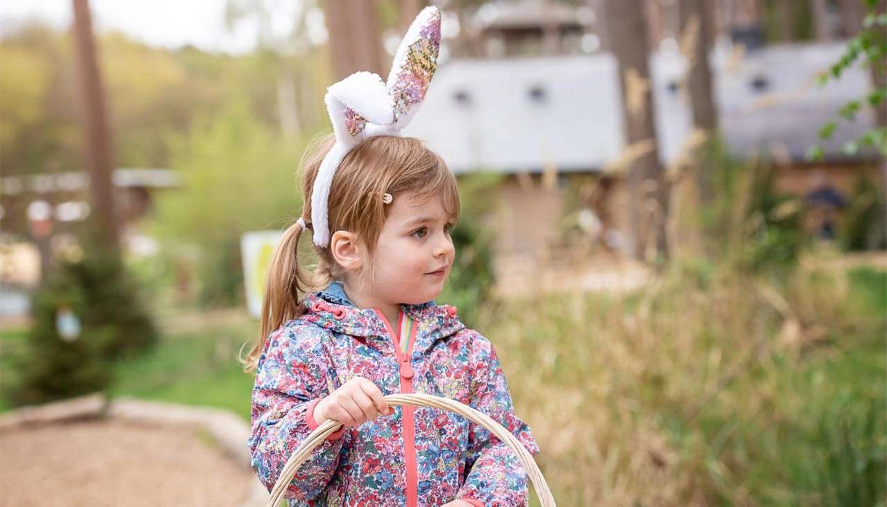 Child wearing sparkly bunny ears grips a wicker basket, standing in a floral raincoat along a garden path in a wooded park.