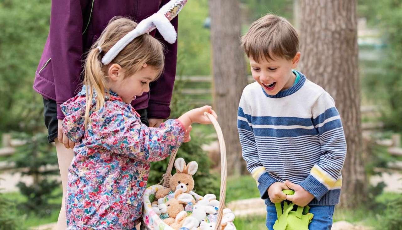 Two children examine a basket of small stuffed bunnies; one wears bunny-ear headband and lifts the handle, the other smiles excitedly. They stand outdoors near trees with an adult nearby.