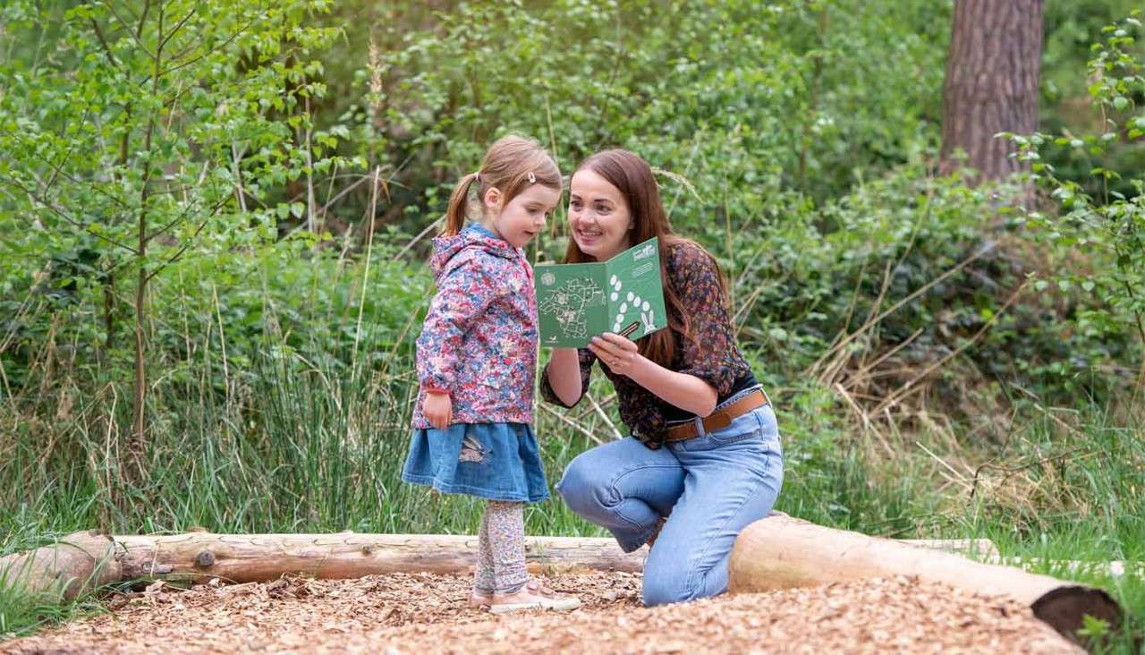 Woman kneeling shows a green booklet to a young girl; both study it together. Outdoors on woodchips beside logs, surrounded by lush green trees and brush in a forest.