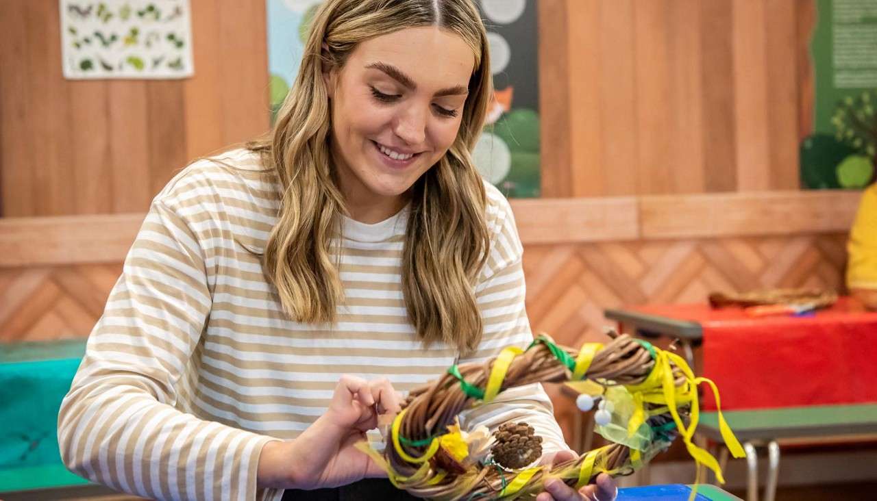 Woman decorates a vine wreath, tying green and yellow ribbons and adding a pinecone, smiling as she works at a craft table in a classroom-like room with posters and supplies.