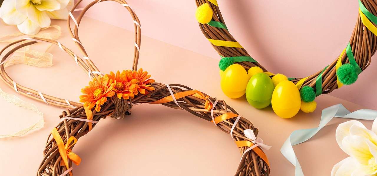 Wicker Easter wreaths sit decorated with orange flowers, ribbons, yellow-green eggs, and pom-poms, resting on a pastel pink-beige surface with scattered ribbons and white blossoms around.