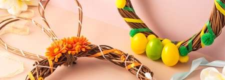 Two grapevine wreaths lie decorated: one bunny-ear shape with orange flowers and striped ribbon; another round with yellow eggs, green pom-poms, and ribbon; set on a pastel pink tabletop.