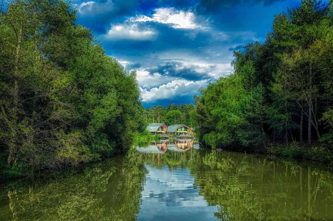 Two curved-roof cabins reflect on a calm river, framed by dense green trees; ripples lead toward them under dramatic clouds in a forested valley.