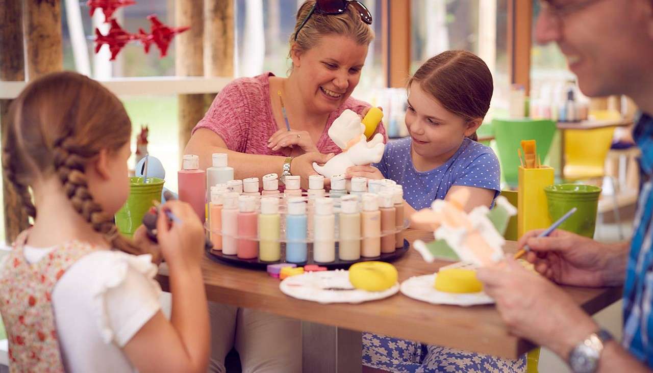 Children and adults paint ceramic figures, dipping brushes into pastel bottles on a rotating tray, in a bright craft studio with wooden posts, big windows, and colorful chairs.