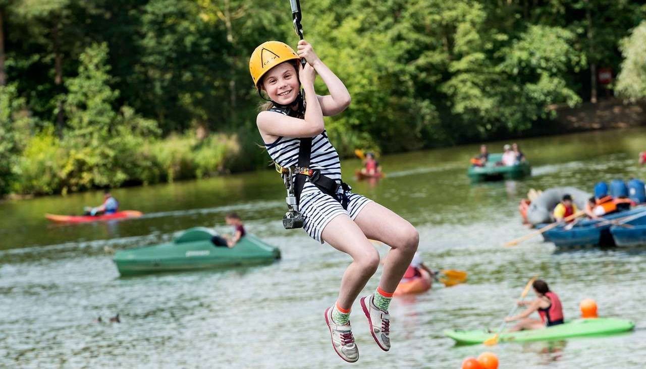 Child wearing helmet and harness ziplines, smiling, above a lake where people paddle kayaks and pedal boats; green trees line the shore on a sunny day.