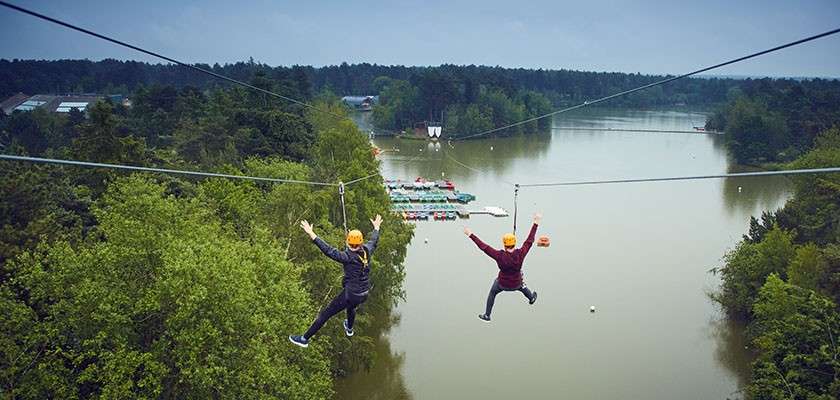 Two helmeted riders zipline side by side, arms outstretched, gliding above a calm lake. Surrounding context: dense forest, docks with moored boats, overcast sky, cables stretching toward distant landing platform.