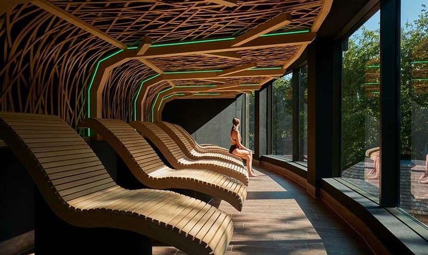 Curved wooden lounge chairs—empty and one occupied—line a windowed relaxation room. A seated person gazes outside, while a lattice ceiling with green accent lighting arches overhead, trees beyond glass.
